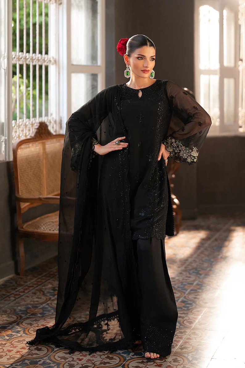 May include: A black ensemble featuring a long-sleeved top with intricate embellishments and wide-leg pants. The outfit includes a sheer shawl and is accessorized with green earrings and a ring. A red flower adorns the hair.