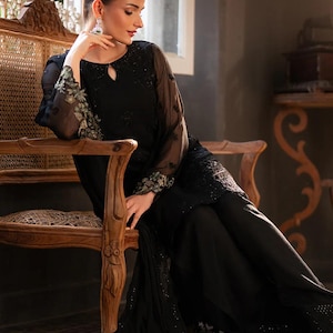 May include: A black formal outfit featuring a long-sleeved top with floral embroidery on the sleeves and a keyhole neckline. The outfit includes matching wide-leg pants. The model is seated on a wooden chair with ornate carvings.