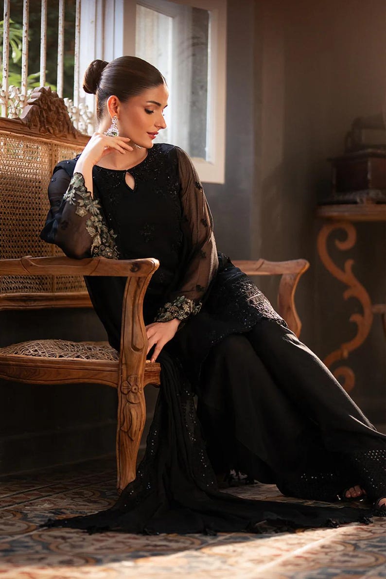 May include: A black formal outfit featuring a long-sleeved top with floral embroidery on the sleeves and a keyhole neckline. The outfit includes matching wide-leg pants. The model is seated on a wooden chair with ornate carvings.