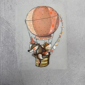 Könnte beinhalten: Eine fantasievolle Illustration eines Dachses und eines Fuchses, die in einem Heißluftballon mit rosa-weißem Streifenmuster fahren. Der Ballon ist mit einer Girlande aus bunten Flaggen geschmückt.