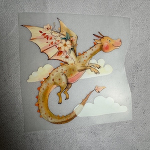 Könnte beinhalten: Eine skurrile Illustration eines fliegenden Drachen mit einer warmen Farbpalette aus Orange, Gelb und Rosa. Die Flügel des Drachen sind mit floralen Akzenten verziert, und er schwebt über flauschigen weißen Wolken. Der Drache hat einen freundlichen Gesichtsausdruck.