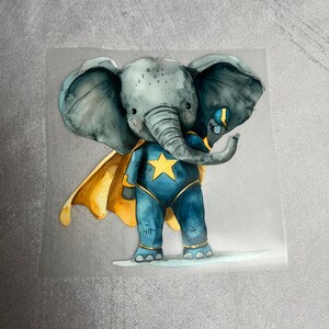 Könnte beinhalten: Eine Aquarell-Illustration eines grauen Elefanten, der ein blaues Superhelden-Kostüm mit einem gelben Umhang und einem Stern auf der Brust trägt. Der Elefant hält einen blauen Ball mit einem gelben Stern in seinem Rüssel.