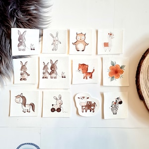 Könnte beinhalten: Eine Sammlung quadratischer, cremefarbener Lederaufnäher mit Aquarellillustrationen verschiedener Tiere, darunter Kaninchen, eine Eule, ein Lama, eine Katze, ein Pferd, eine Kuh und ein Zebra. Die Aufnäher sind auf einer weißen Oberfläche angeordnet.