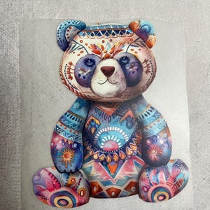 Könnte beinhalten: Ein farbenfroher, gemusterter Teddybär mit einem blauen, rosa und orangenen Design. Der Bär hat ein weißes Gesicht mit schwarzen Augen und einer schwarzen Nase.