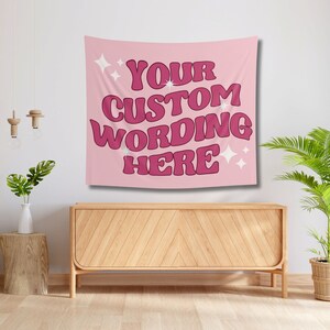 May include: A pink wall tapestry with the text "YOUR CUSTOM WORDING HERE" in a retro font. White starburst graphics are on the top left and bottom right. The tapestry is hanging above a wooden cabinet.