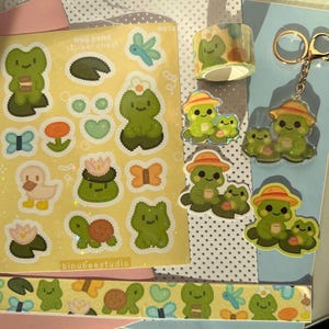 Könnte beinhalten: Eine Sammlung von Schreibwaren und Accessoires zum Thema Frosch. Enthält ein Stickerbogen mit verschiedenen Froschmotiven, eine Rolle Washi-Tape, einen Schlüsselanhänger und mehrere froschförmige Aufkleber. Die Artikel sind in Grün-, Gelb- und Orangetönen gehalten.