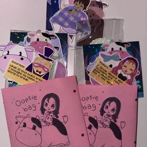 Könnte beinhalten: Eine Sammlung handgefertigter Aufkleber und kleiner rosa Taschen. Die Aufkleber zeigen Cartoon-Kühe, Menschen und Textblasen mit Sätzen wie "Gourmet" und "Popuri". Die rosa Taschen tragen den Text "Oopsie bag" und eine Zeichnung eines Mädchens, das auf einer Kuh reitet.