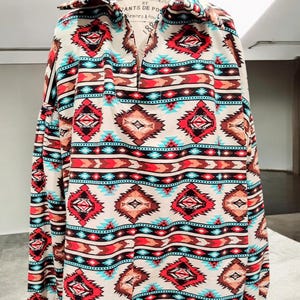 Puede incluir: Una camisa blanca de manga larga con un estampado geométrico colorido. El estampado presenta colores rojo, azul, marrón y negro. La camisa tiene cuello y cierre de botones en la parte delantera.