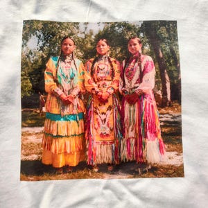 May include: White t-shirt featuring a printed image of three women in traditional Native American dresses. The dresses are brightly colored with intricate beadwork and fringe. The women stand in front of a blurred background of trees.