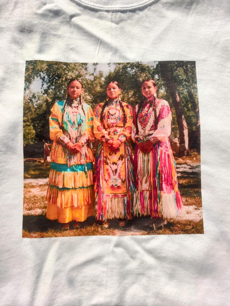 May include: White t-shirt featuring a printed image of three women in traditional Native American dresses. The dresses are brightly colored with intricate beadwork and fringe. The women stand in front of a blurred background of trees.
