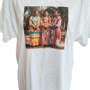 May include: White t-shirt featuring a printed image of three women in traditional Native American attire. The women are standing in front of a blurred background of trees. The women are wearing colorful dresses with intricate patterns.
