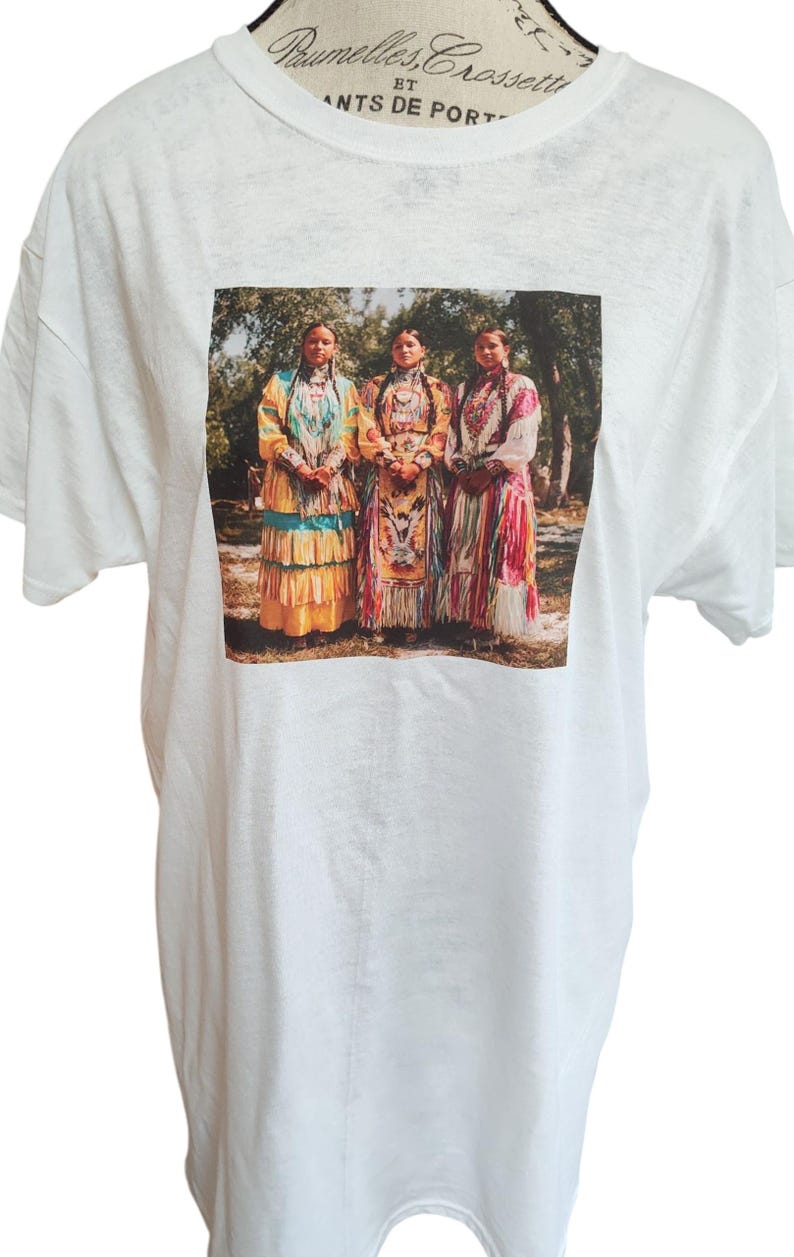 May include: White t-shirt featuring a printed image of three women in traditional Native American attire. The women are standing in front of a blurred background of trees. The women are wearing colorful dresses with intricate patterns.