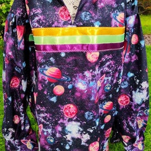May include: A long-sleeved shirt with a black background and a galaxy print featuring planets and stars. The shirt has a collar and a band of yellow, green, and purple ribbon around the waist. The text "Bumelles, Crosse ET TIRANTS DE PORTE Yous Genres & tous Style! S 1889" is printed on the collar.