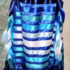 May include: A blue, white, and turquoise ribbon skirt with long, flowing ribbons. The skirt is made of multiple layers of ribbon sewn together to create a striped pattern. The ribbons are tied at the waist and flow down to the floor.
