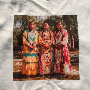 May include: White t-shirt featuring a printed image of three women in traditional Native American attire. The women are standing in front of a blurred background of trees. Their dresses are colorful, with intricate beadwork and patterns.
