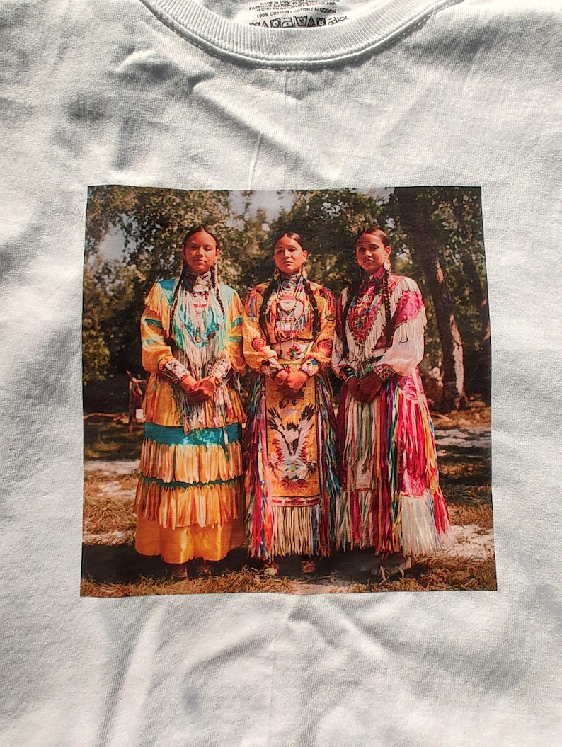 May include: White t-shirt featuring a printed image of three women in traditional Native American attire. The women are standing in front of a blurred background of trees. Their dresses are colorful, with intricate beadwork and patterns.