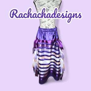 May include: A purple satin skirt with a striped pattern of purple, white and beige ribbon. The skirt has a gathered waistband and is decorated with long, flowing ribbons.