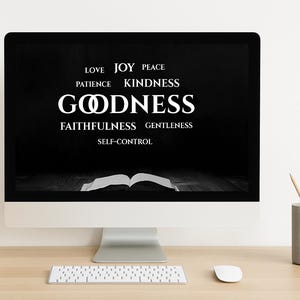 Può includere: Un computer mostra la parola "GOODNESS" in bianco su sfondo nero, circondata da parole come "LOVE" e "JOY". Una tastiera e un mouse bianchi sono su una scrivania di legno, con una pianta e un portapenne.