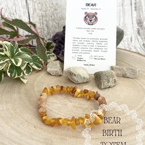 May include: A stretch bracelet made of irregular amber chips and round wooden beads. Behind it, a white card displays the text BEAR, August 22 - September 21, with a bear illustration and descriptive text about the bear totem and birth stones.