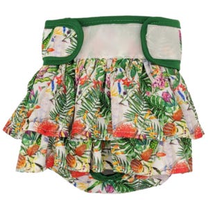 Puede incluir: Un pañal para perro verde y blanco con un estampado tropical con flores, hojas y pájaros. El pañal tiene una cintura elástica verde y un cierre de velcro blanco.