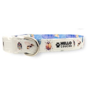 Könnte beinhalten: Weißes Hundehalsband mit einem blau-weißen Muster aus Bienen und Käfern. Das Halsband hat eine silberne Schnalle und ein schwarzes Etikett mit der Aufschrift "HELLO COUCOU" und einem Pfotenabdruck.