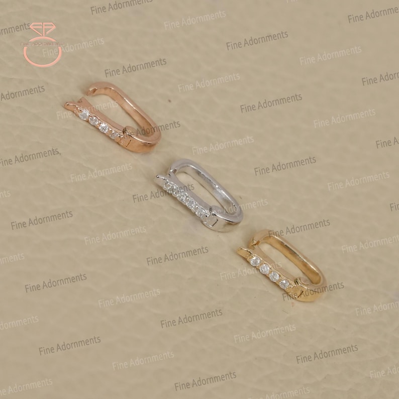 May include: Three rectangular jewelry clasps in rose gold, silver, and gold. Each clasp features a row of small, clear gemstones. The clasps are displayed on a neutral, textured surface.