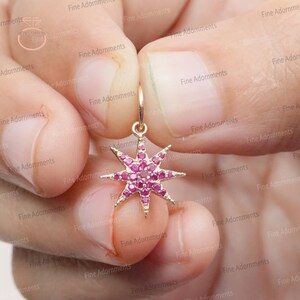 May include: A gold-coloured starburst earring with a hoop. The starburst is encrusted with small, round, pink gemstones. The earring is held between fingers, showcasing the jewellery's design and colour.