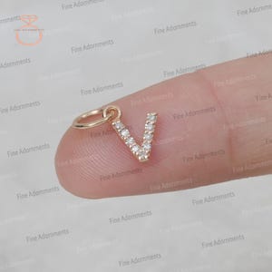 Pode incluir: Um amuleto de ouro em forma da letra V, com pequenos diamantes cravejados na letra. O amuleto está em um dedo.