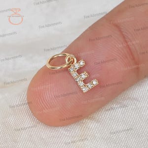 以下が含まれることがあります： 小さな透明な宝石がちりばめられた、金色の文字「E」の形をしたチャーム。チャームは小さなリングに付いています。チャームは指で持たれており、背景はぼやけています。