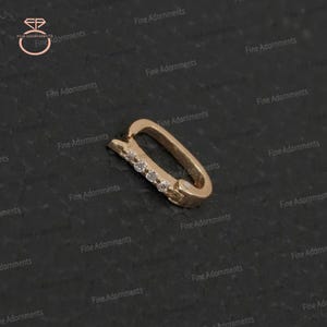May include: A gold-colored, rectangular earring clasp with a row of small, clear gemstones. The clasp is slightly curved and has a hinged design. The background is a dark, textured surface.