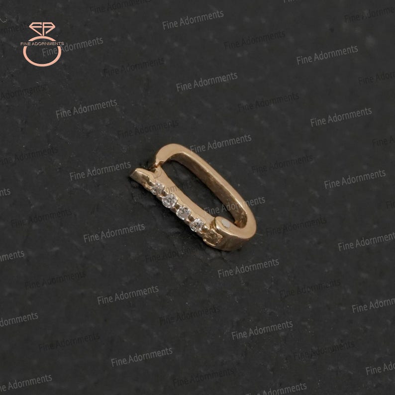 May include: A gold-colored, rectangular earring clasp with a row of small, clear gemstones. The clasp is slightly curved and has a hinged design. The background is a dark, textured surface.