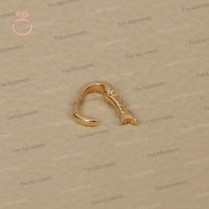 May include: A gold-colored nose ring with a curved design and a small decorative element. The nose ring is made of metal and has a smooth, polished finish. The jewelry is designed to be worn in the nose.
