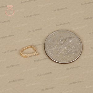 May include: A gold-colored jewelry clasp with small, clear stones set along one edge. The clasp is next to a US dime for size comparison. The clasp has a curved, oval shape.