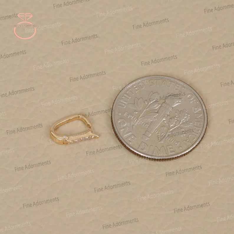May include: A gold-colored jewelry clasp with small, clear stones set along one edge. The clasp is next to a US dime for size comparison. The clasp has a curved, oval shape.