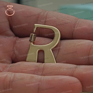 Pode incluir: Brinco de letra R em tom dourado. O brinco é uma única letra R com um poste e uma parte de trás.