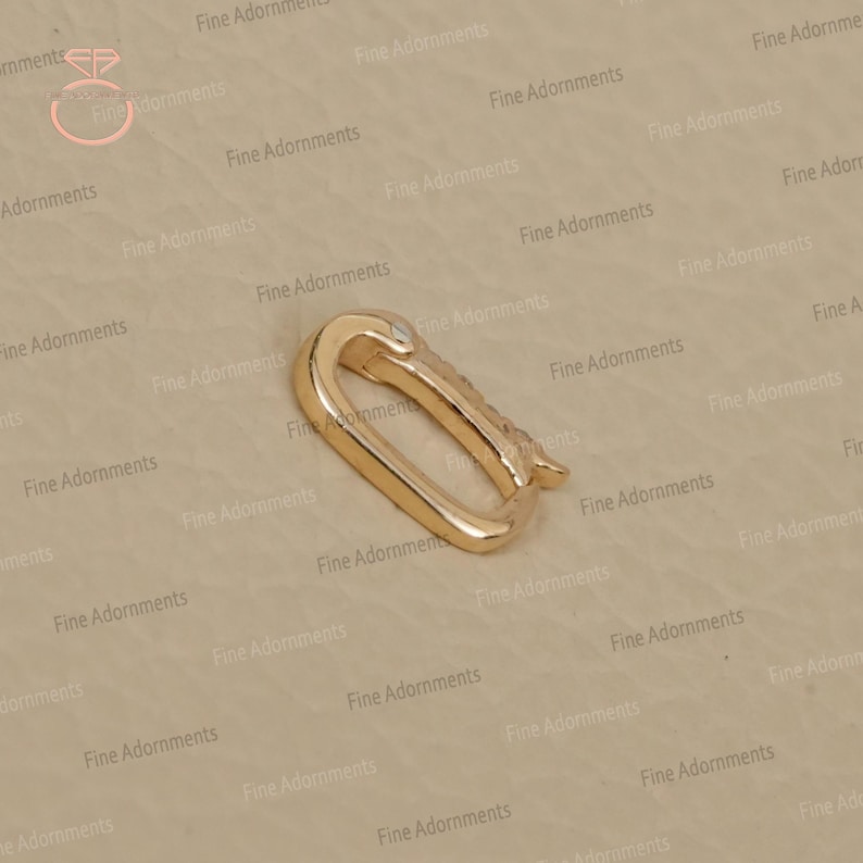 May include: A gold-colored, oval-shaped clasp with a hinged closure. The clasp is made of a shiny metal and has a small, decorative element on the side. The background is a neutral beige color.