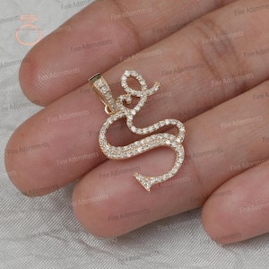 Puede incluir: Un colgante dorado con forma de la letra "S", incrustado con pequeños diamantes brillantes. El colgante tiene un lazo en la parte superior para una cadena. El diseño es elegante y adecuado para joyería.
