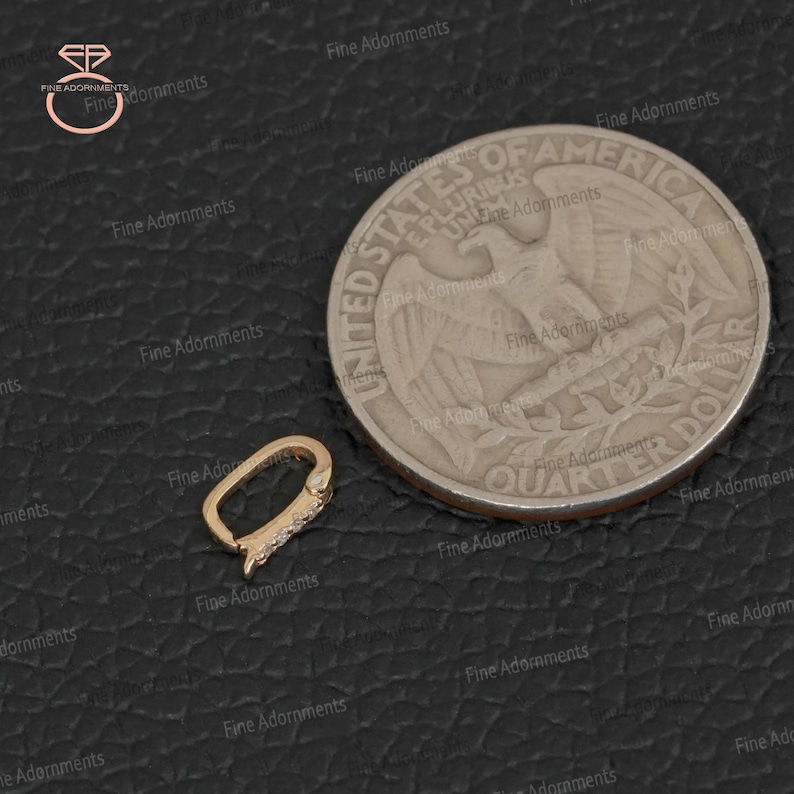 May include: A gold-colored oval clasp with small, clear stones, next to a United States quarter. The clasp is open, showing its design. The background is a dark, textured surface.