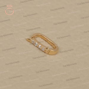 May include: A gold-colored clasp with a rectangular shape and a row of five small, clear gemstones. The clasp is designed to open and close, likely for jewelry or accessories. The background is a light beige color.
