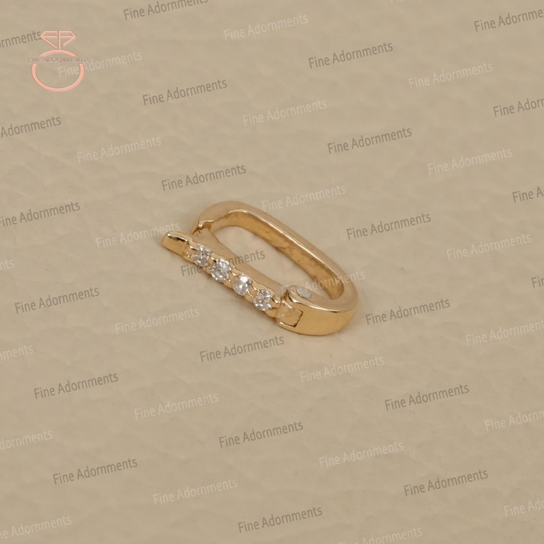 May include: A gold-colored clasp with a rectangular shape and a row of five small, clear gemstones. The clasp is designed to open and close, likely for jewelry or accessories. The background is a light beige color.