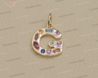 以下が含まれることがあります： ベージュの背景に、Gの文字の形をした金色のチャーム。カラフルな宝石で飾られています。