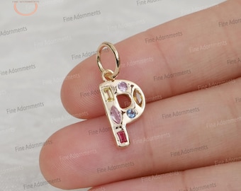 以下が含まれることがあります： 様々な色の宝石がちりばめられた、金色の文字「P」のペンダント。ペンダントの上部にはチェーンを通すためのループが付いています。宝石はピンク、黄色、赤など、様々な形と色をしています。