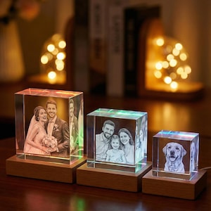 May include: Three illuminated crystal photo blocks with wooden bases. The blocks display engraved images of a couple, a family, and a dog. Each block emits a soft, colourful glow, creating a warm ambience. The blocks are on a dark wooden surface.