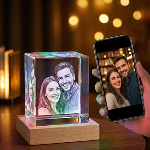 Puede incluir: Un bloque de cristal rectangular transparente con una foto de una pareja sonriente, iluminada con luces de colores. El bloque se encuentra sobre una base de madera. Un smartphone muestra la misma foto.
