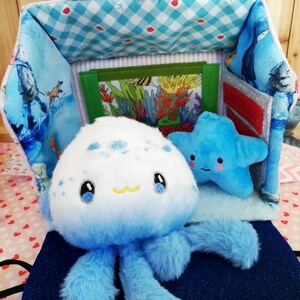 Può includere: Un giocattolo di peluche a forma di medusa con testa bianca e tentacoli blu, accanto a un giocattolo a forma di stella marina blu. I giocattoli sono davanti a una borsa di stoffa con un motivo a quadri blu e bianchi e una colorata scena sottomarina.