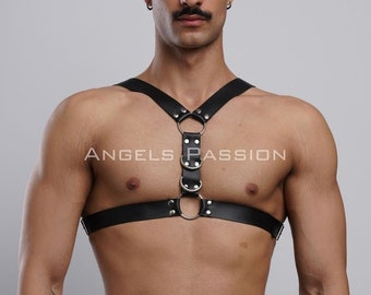 May include: A black leather harness with silver metal rings and studs. The harness has two straps that go over the shoulders and connect in the front with a buckle. The harness is worn by a person with a mustache.