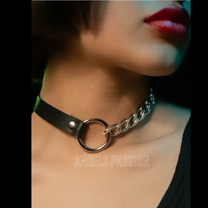 Può includere: Un collare di pelle nera con una catena argentata e un grande anello argentato. Il testo "Angels Passion" è stampato nella parte inferiore dell'immagine.