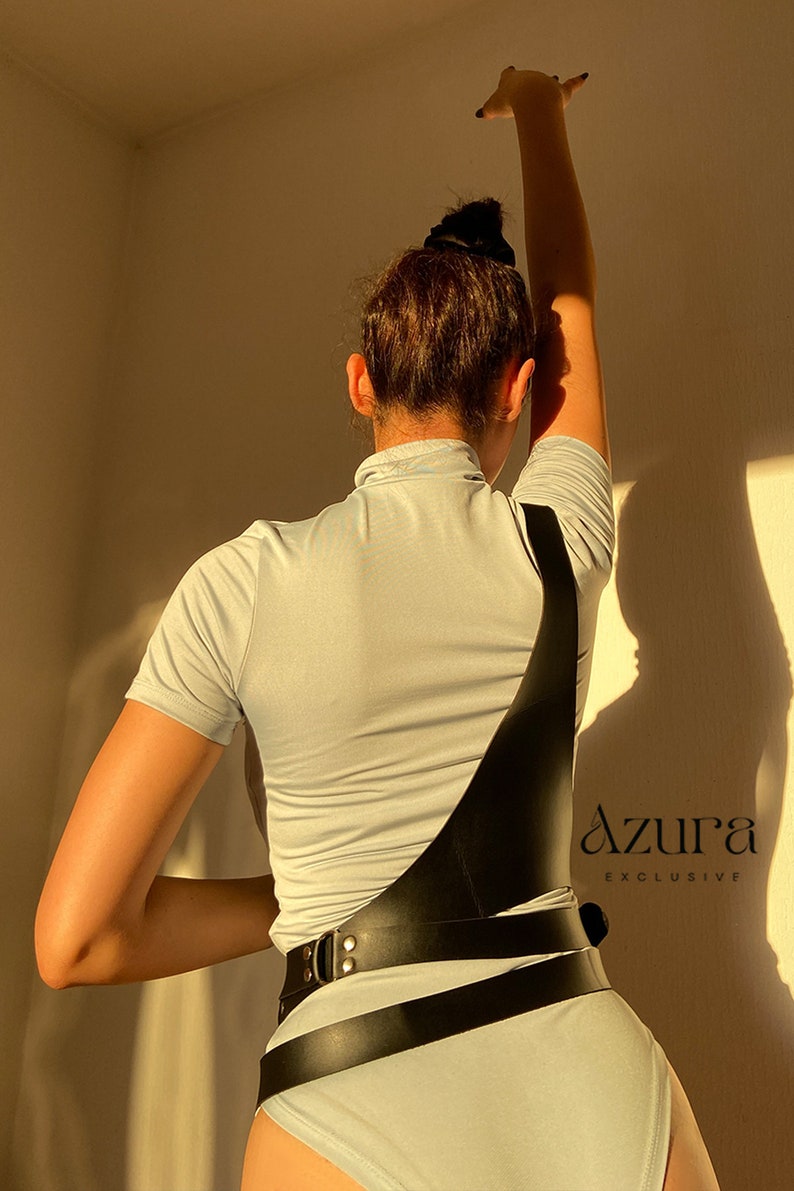Puede incluir: Un arn&eacute;s de cuerpo de cuero negro con cierre de hebilla y dise&ntilde;o de correas. El arn&eacute;s se lleva sobre una camiseta de manga corta azul claro y lleva el texto "Azura Exclusive".