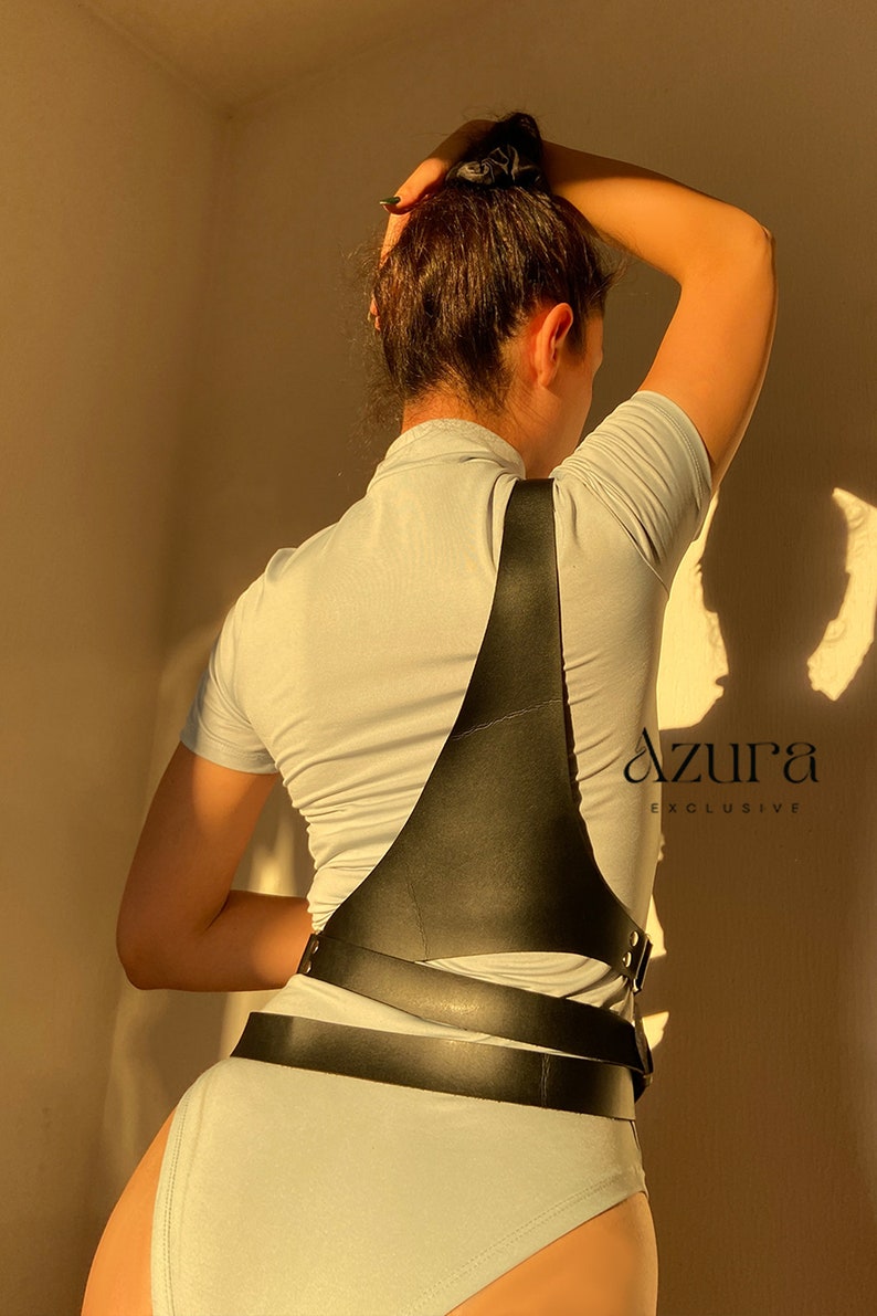 Puede incluir: Arn&eacute;s de cuerpo de cuero negro con correas ajustables y herrajes met&aacute;licos. El arn&eacute;s se lleva sobre un body azul claro. El texto "AZURA EXCLUSIVE" es visible en el lado derecho de la imagen.
