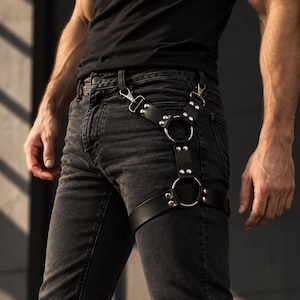 May include: A black leather leg harness featuring two silver-toned metal rings connected by adjustable straps with silver rivets. The harness attaches to belt loops with two metal clips and secures around the thigh with a wide leather strap.